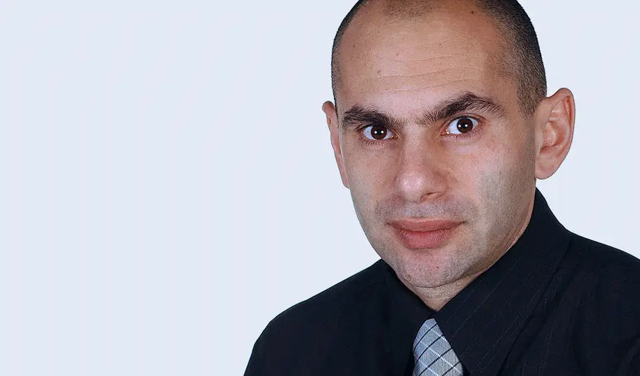 Portrait of Professor Costas Karageorghis, man in black suit with striped tie on light background.