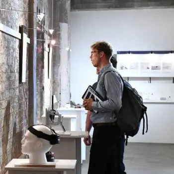 Person standing in an art gallery looking at exhibits with a backpack and holding books.