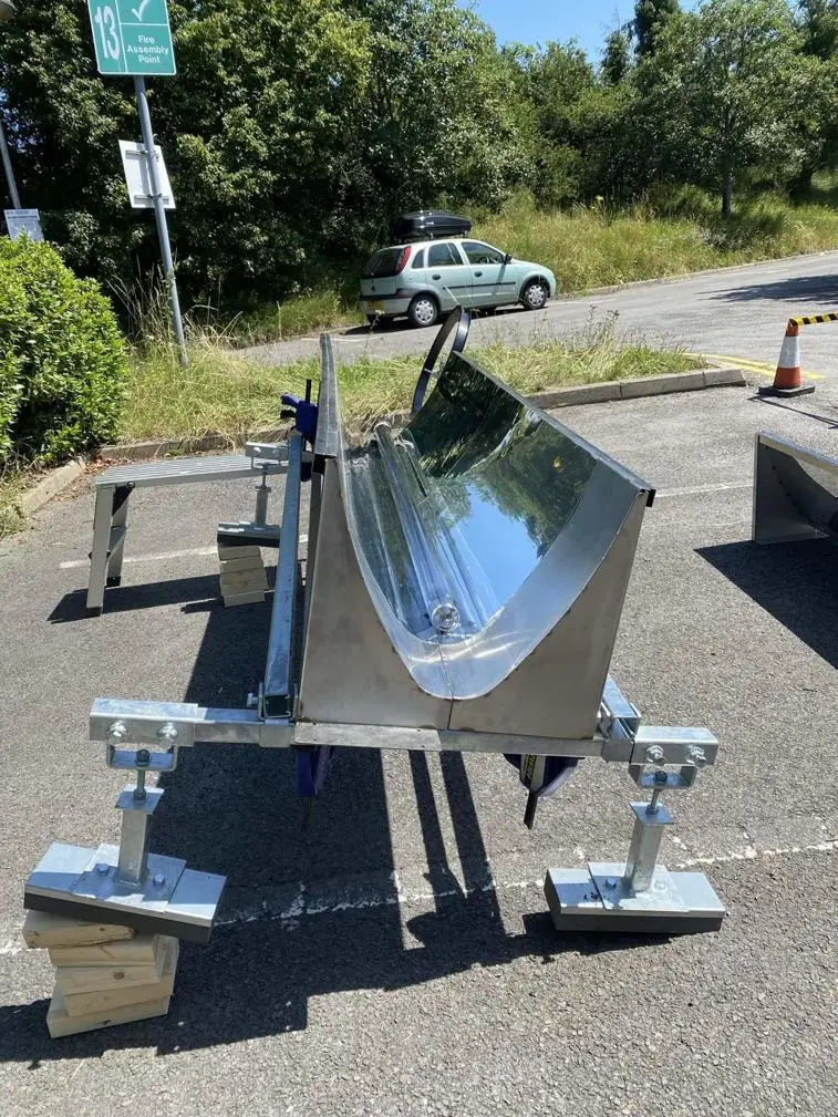 Metal trough on stands, reflecting sunlight, beside a car in a grassy area.