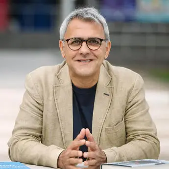 Portrait photo of Professor Daljit Nagra, a man in beige jacket with clasped hands sitting at a table outdoors.