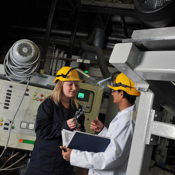 Two industrial workers with hard hats discussing over a clipboard in a factory setting.