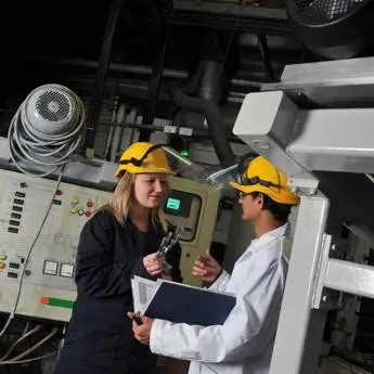 Two industrial workers with hard hats discussing over a clipboard in a factory setting.