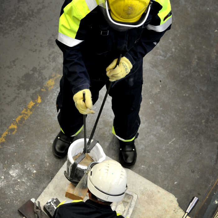 Worker in high-visibility clothing using tongs to handle material in industrial setting.