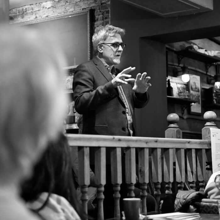 Person gesturing while speaking in a room with listeners. Black and white photo.