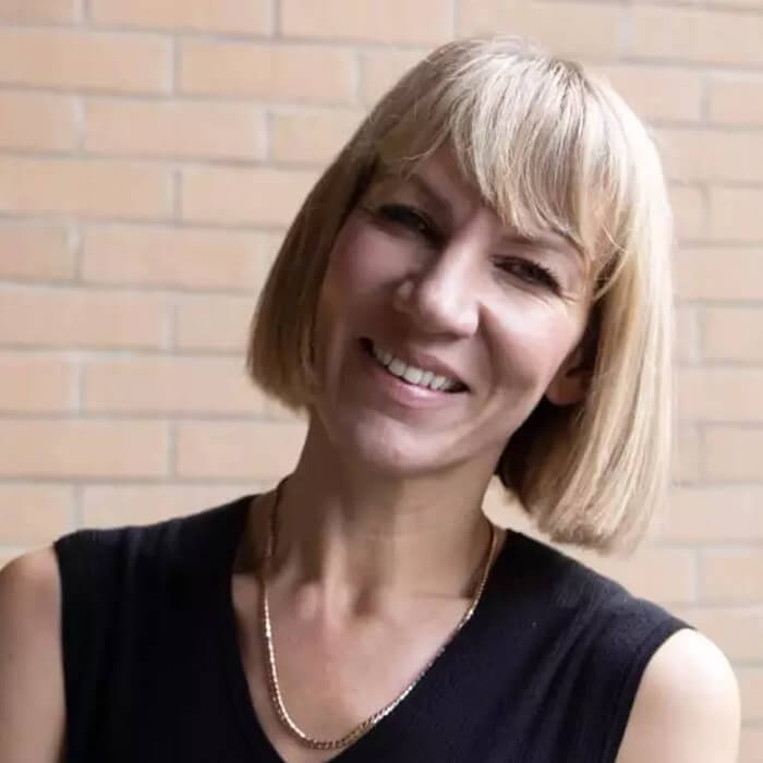 Person with blonde bob haircut wearing a black top and a necklace against a brick wall.