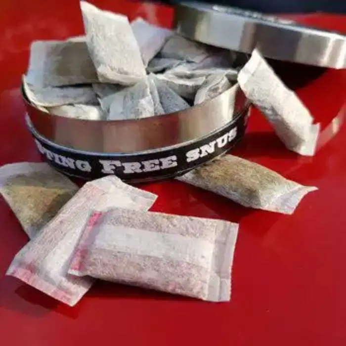 Metal tin labelled "STRONG FREE SNUUS" with open lid and nicotine pouches on a red surface.