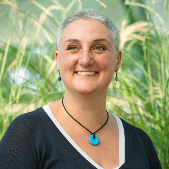 Portrait photo of Dr Lorna Anguilano, wearing a black shirt with white trim and a blue pendant necklace against a green plant background