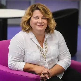 portrait photo of Professor Alexandra Xanthaki, a woman in white shirt with gold necklace sitting on a purple sofa.