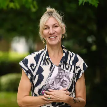 Portrait photo of Professor Hannah Lowe, a woman in a patterned shirt holding a book with a vintage photo cover.