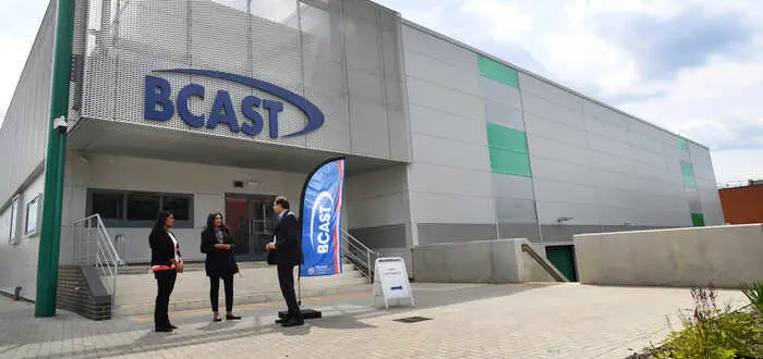 Three people conversing outside a modern building with a BCAST logo above the building entrance.