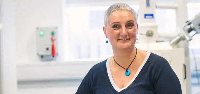 portrait photo of Dr Lorna Anguilano, a person in a lab with a V-neck shirt and a turquoise pendant, standing near machinery.