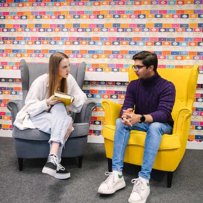 Pair of students discussing a book whilst sitting on arm chairs