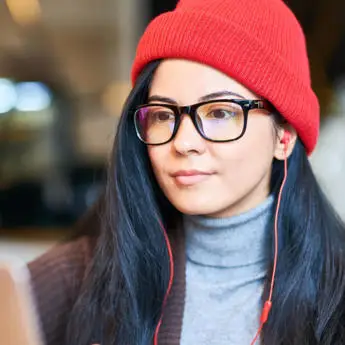 Person wearing a red beanie and earphones with a grey turtleneck and brown vest.