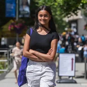 Woman in black top and white pants standing with arms crossed on a sunny campus concourse