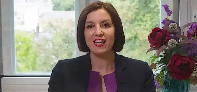 Woman in a purple blouse and black blazer, sitting indoors near a window with flowers